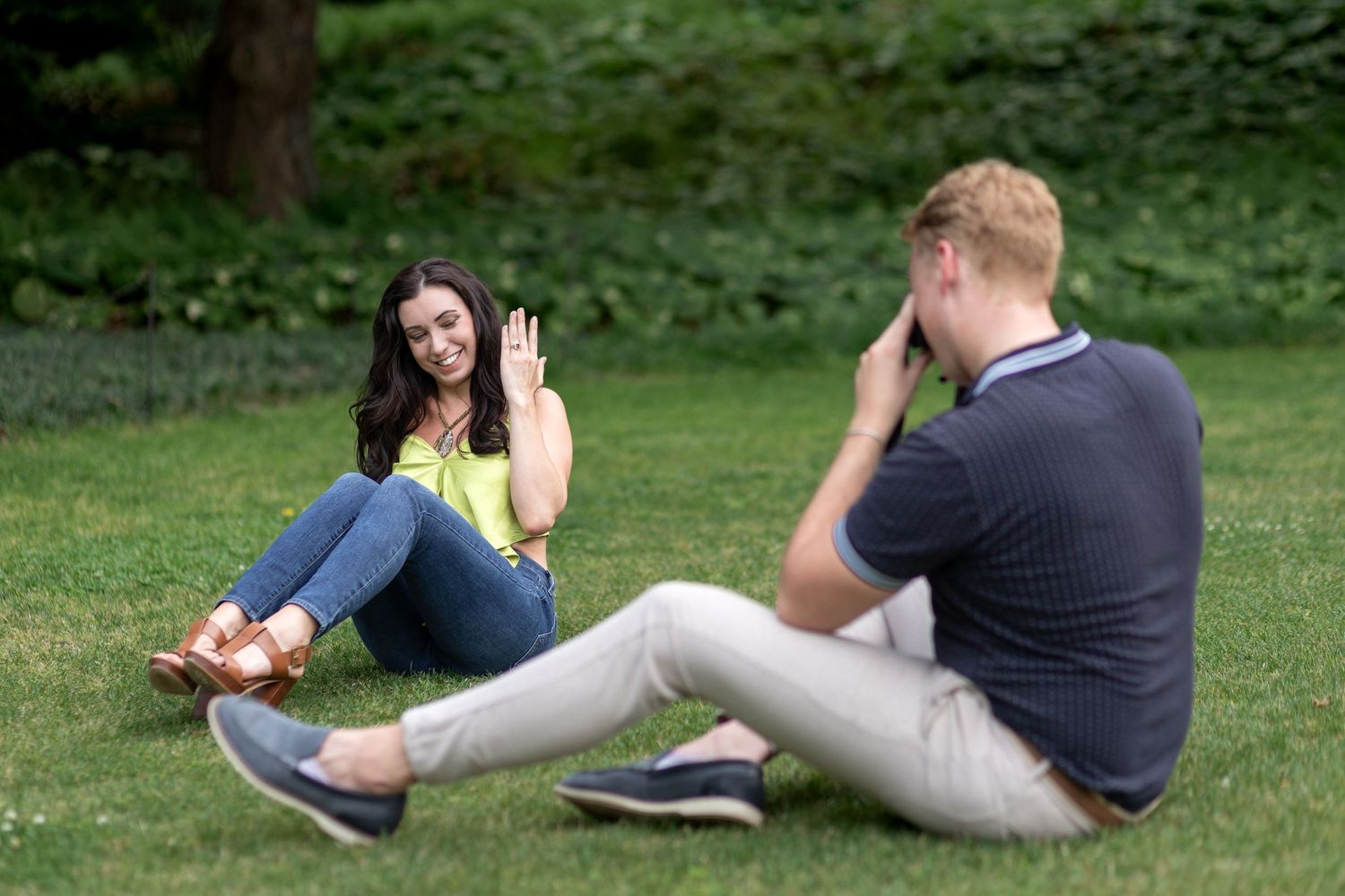 A woman waves at a man photographing her while sitting on green grass in a park.