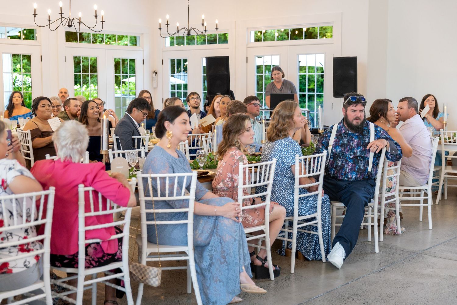Wedding guests seated on white chairs in a bright reception hall with chandeliers and large windows, watching a ceremony or toast.