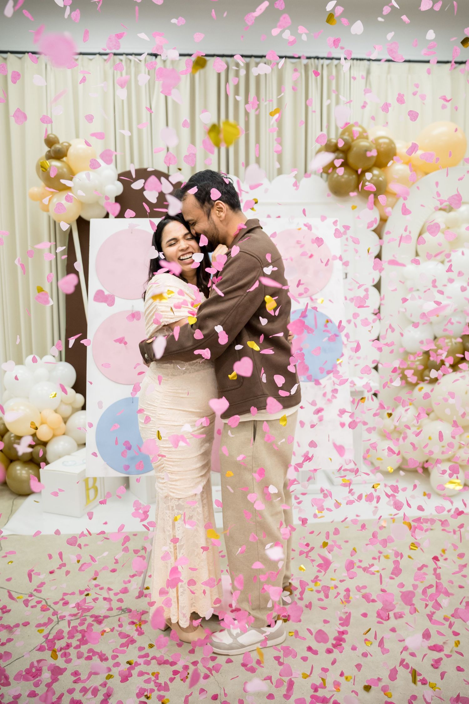Couple embracing joyfully surrounded by falling pink confetti at a gender reveal party celebration.