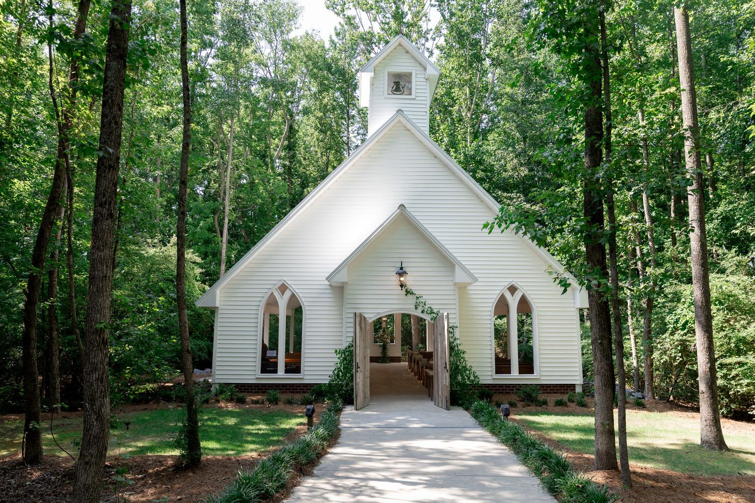 White wooden wedding chapel with arched windows and a steeple, surrounded by tall green trees along a stone pathway.