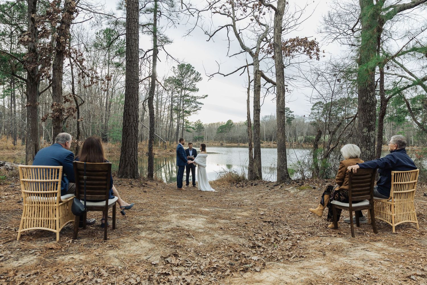 Intimate outdoor lakeside wedding ceremony with a couple exchanging vows and four guests seated in wicker chairs among tall pine trees.