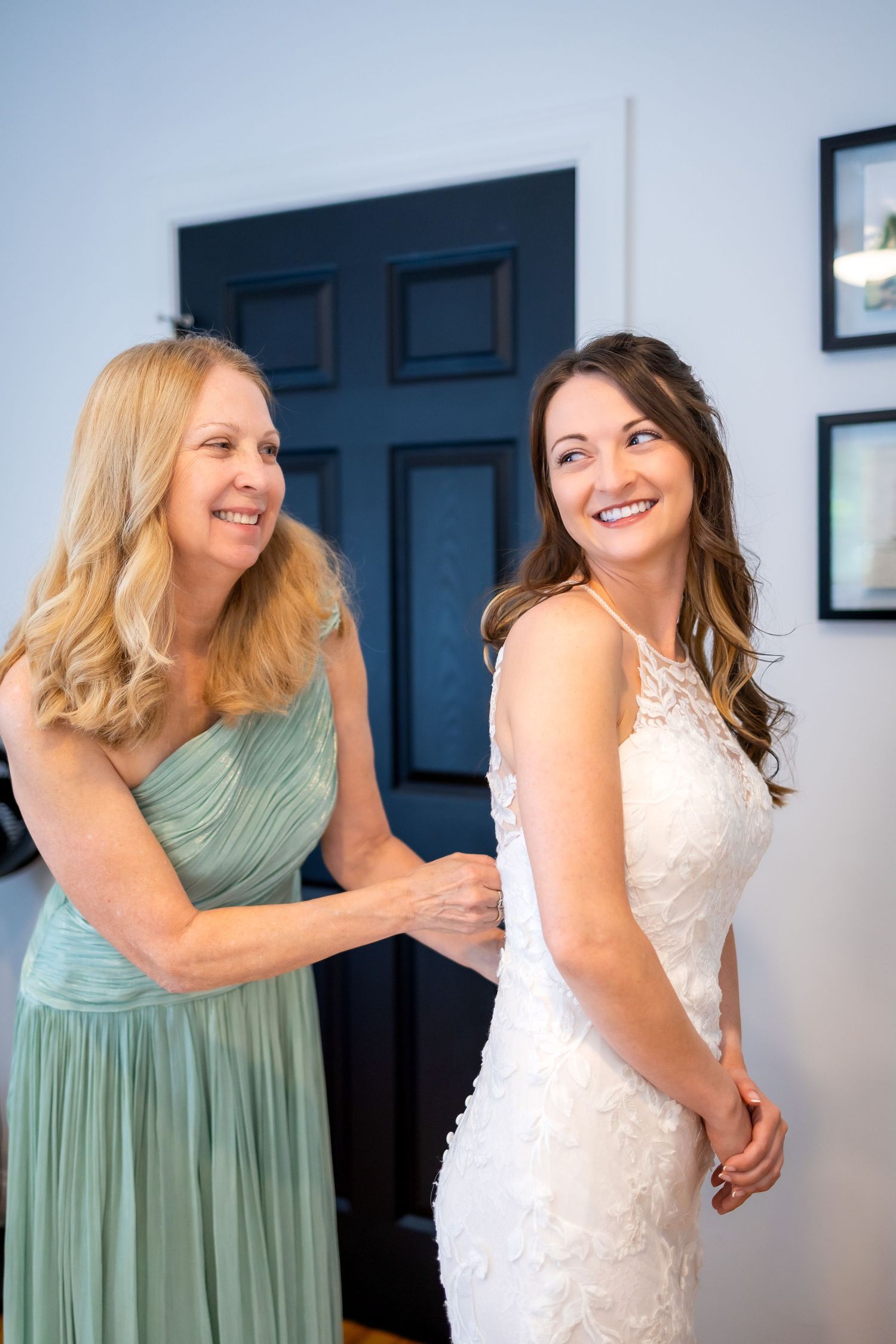 A blonde woman in teal dress helping a smiling bride button her white lace wedding gown.