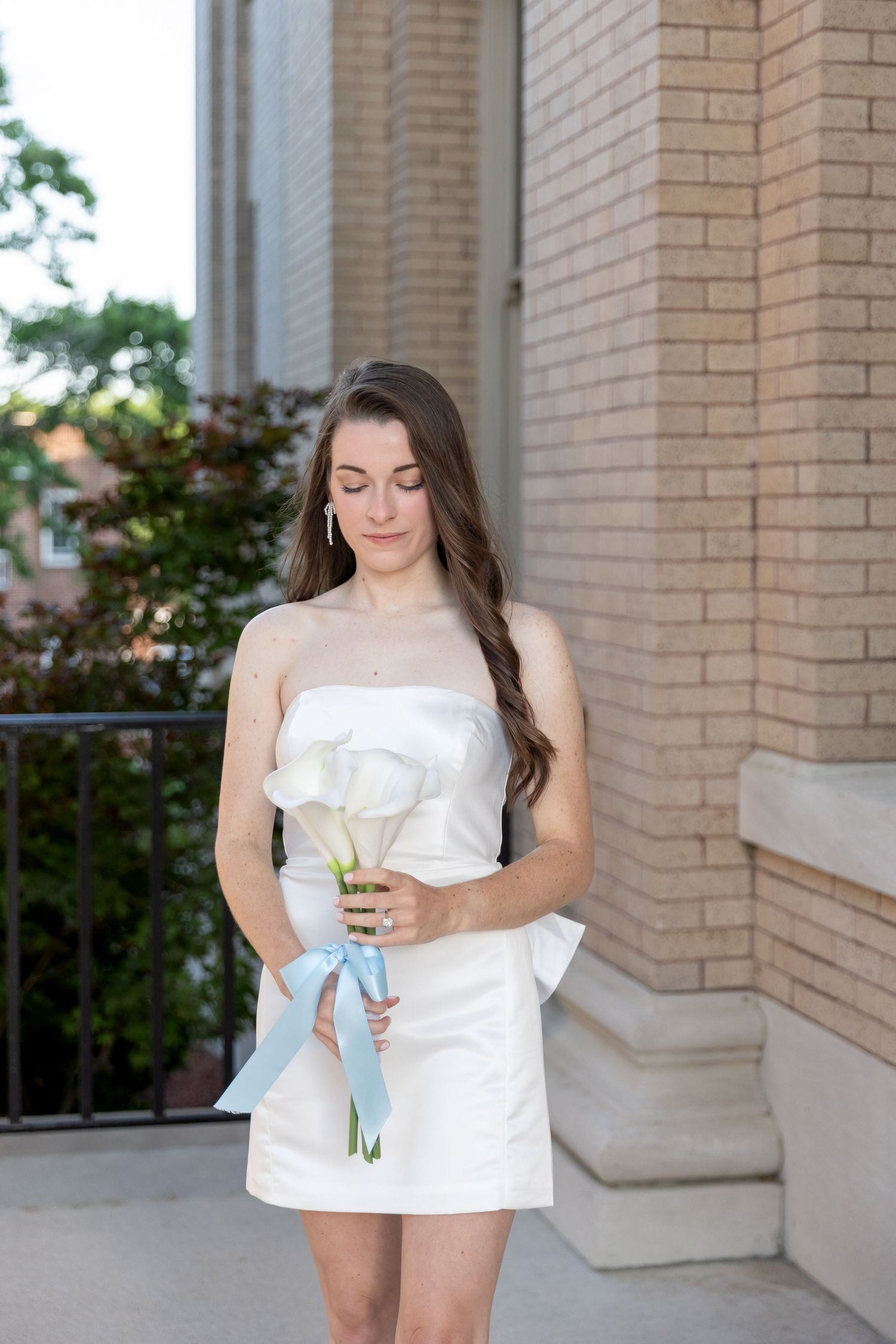 Elegant bride in a white strapless mini dress holds white calla lilies with a blue ribbon outdoors.
