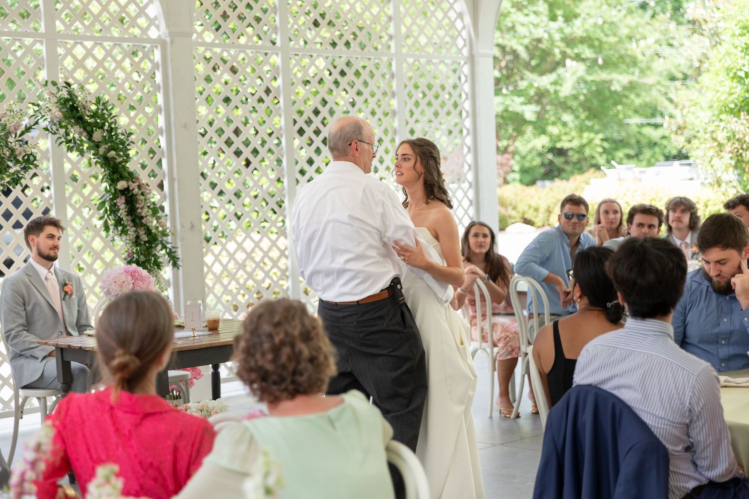 Bride in white gown dances with her father under a white arched pavilion at an outdoor wedding reception.