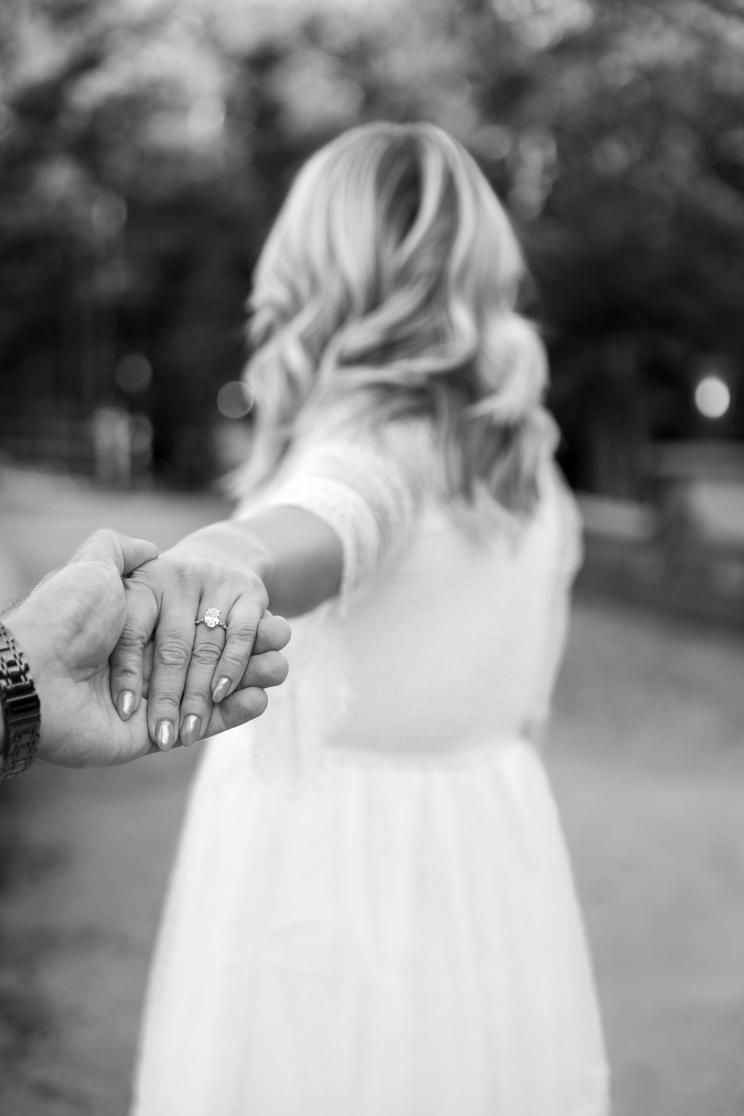 Black and white photo of a woman in a white dress leading her partner by the hand, her engagement ring visible, along a tree-lined path.