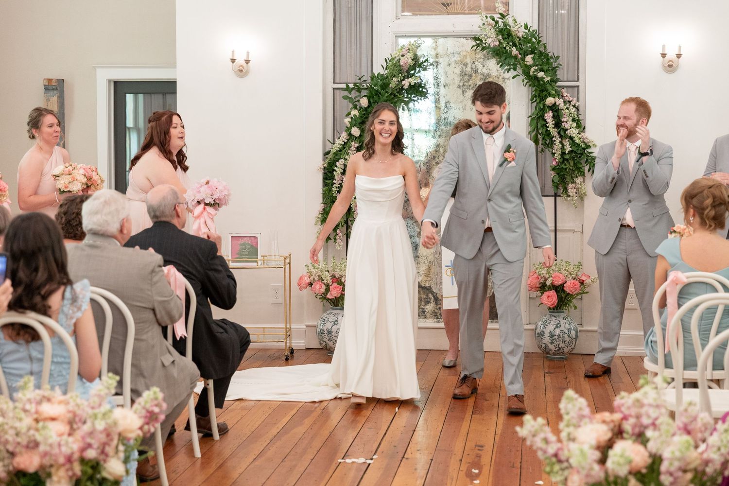 Smiling bride in a white gown and groom in a gray suit hold hands in a floral-decorated aisle during their wedding ceremony as guests watch.