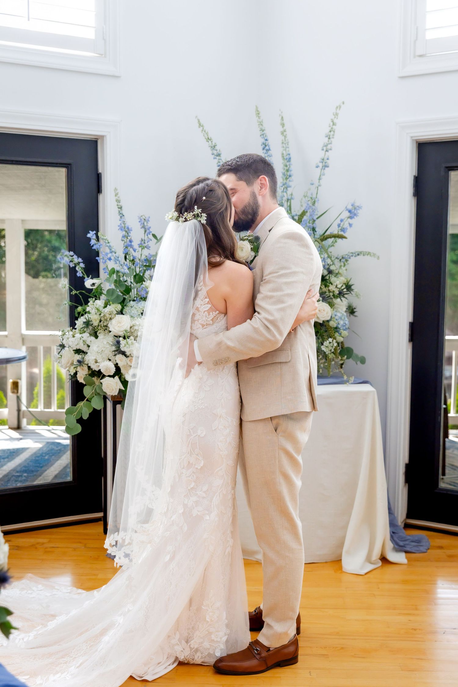 Bride in a lace wedding gown and floral hair piece embraces groom in a tan suit during a ceremony beside blue and white flower arrangements.