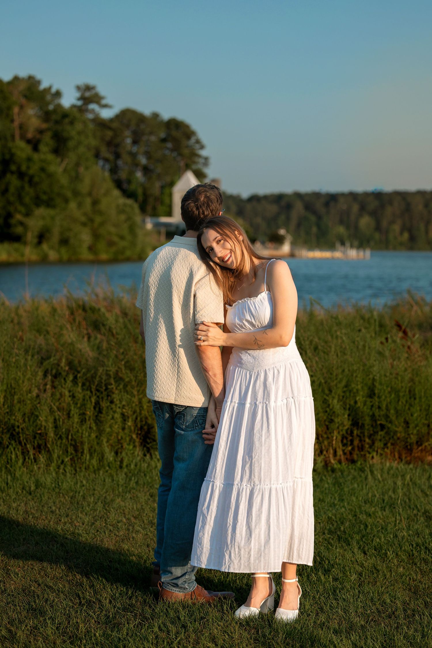 Couple embracing outdoors near a lake at golden hour, woman in white dress smiling.