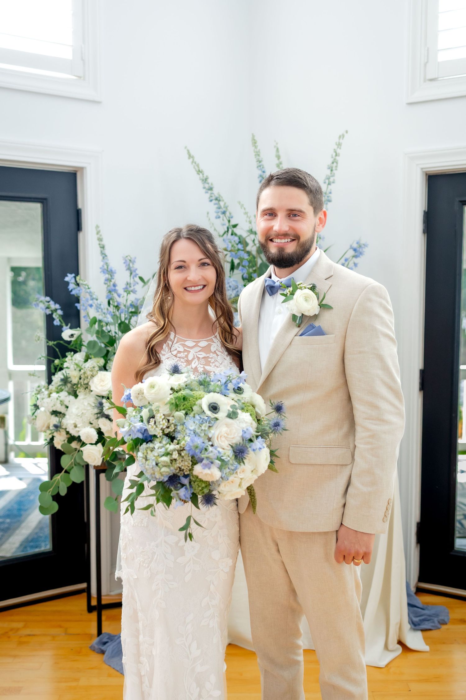 A smiling bride and groom in a tan suit pose with blue and white bouquets in a bright room.
