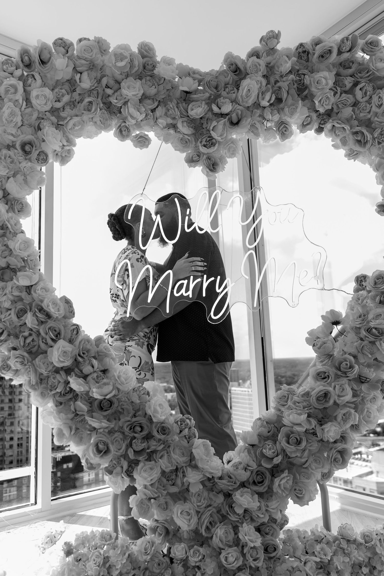 Black and white photo of couple embracing inside a floral heart arch with Will You Marry Me sign.