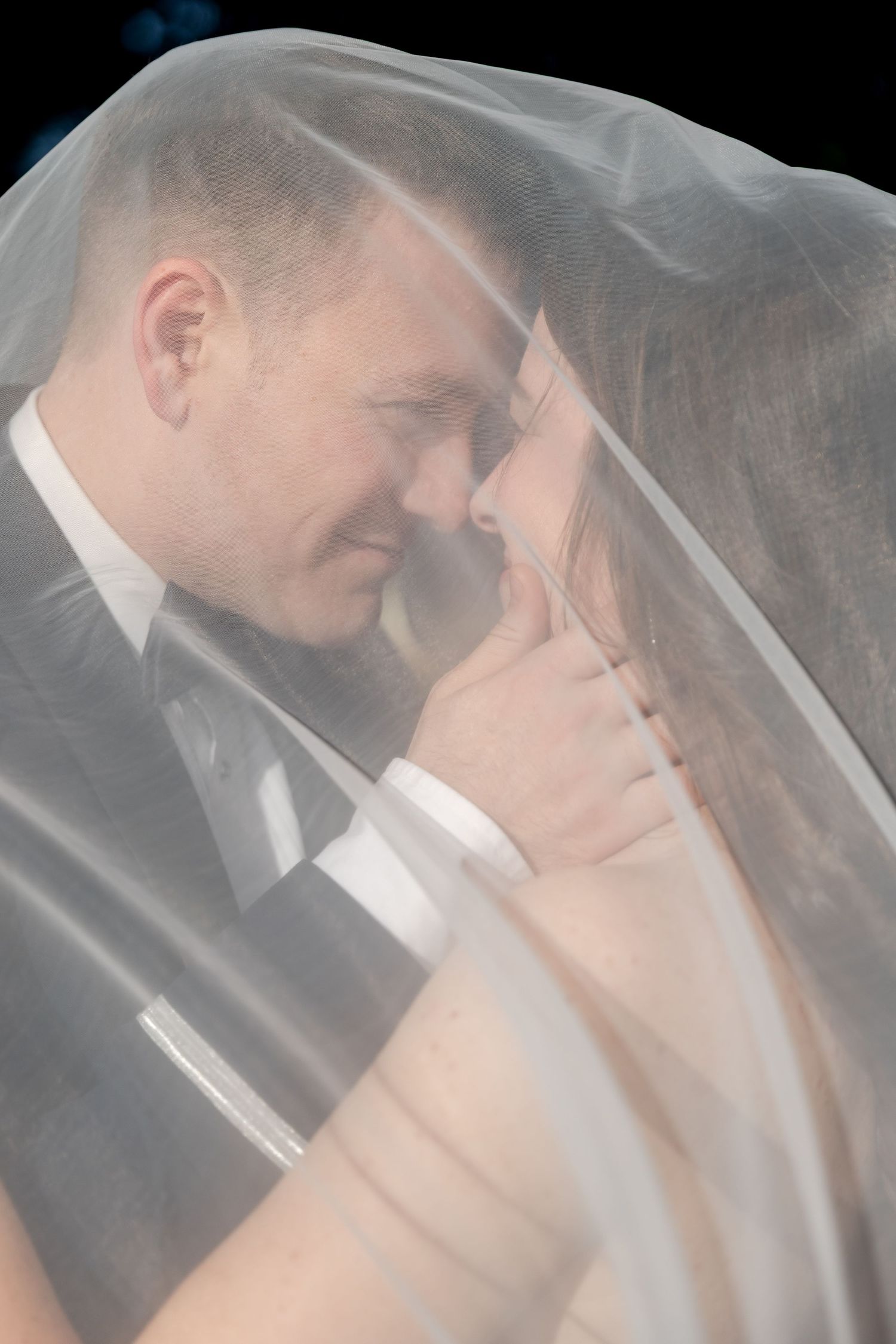 Bride and groom sharing a romantic moment under a sheer wedding veil against a dark background.
