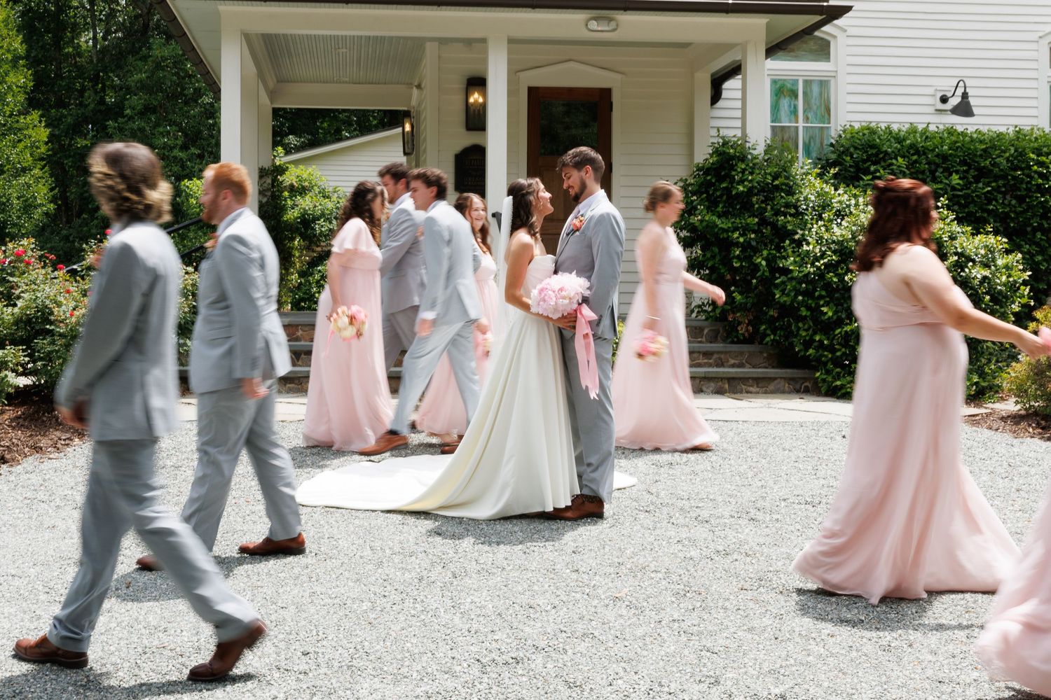 Wedding party in pink and gray attire walks outside white chapel surrounded by lush greenery.