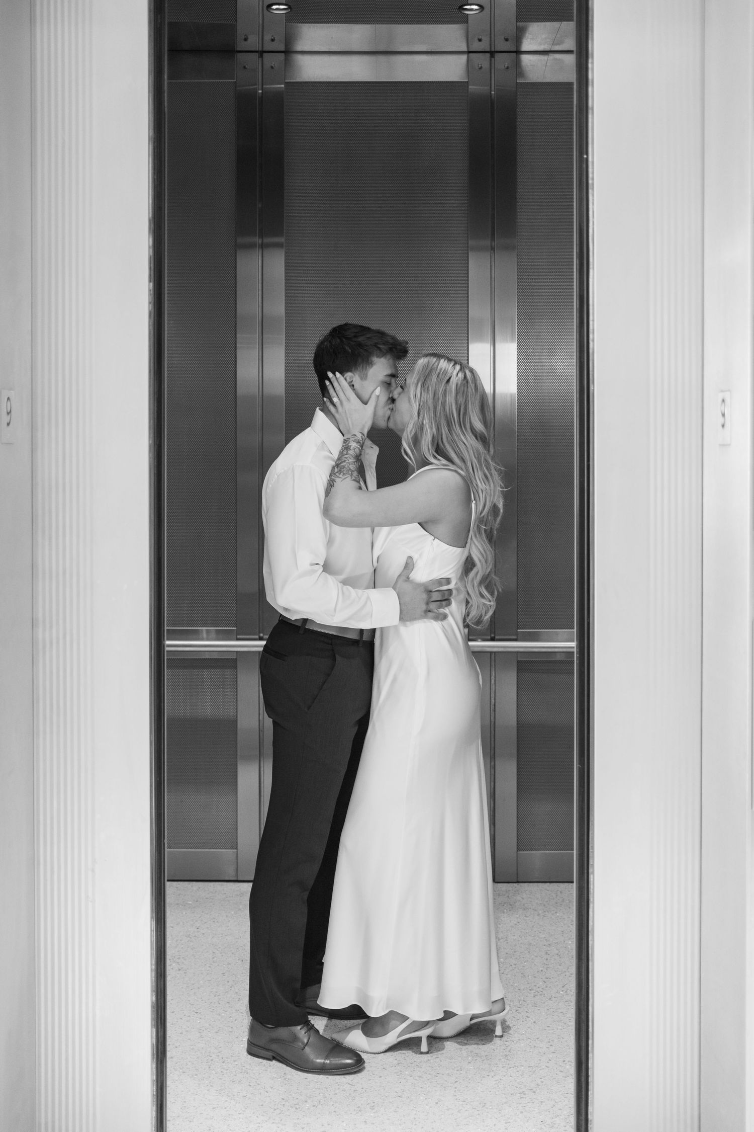 Black and white photo of a couple kissing in an elevator, bride in white gown and groom in dress shirt and trousers.Wake country courthouse