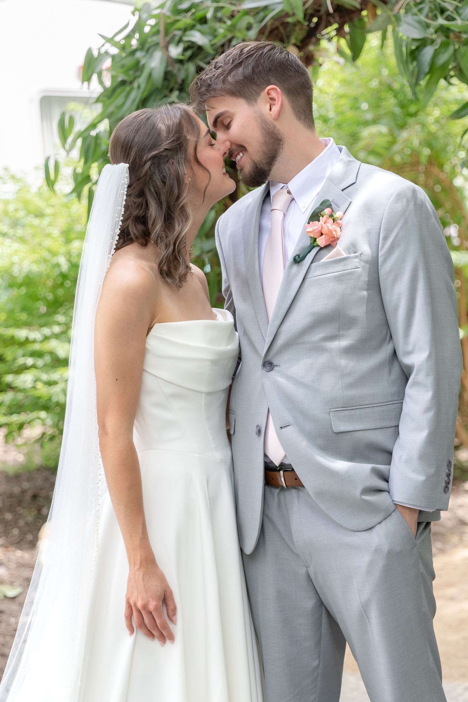 Bride in a strapless white gown and veil kisses groom in a light gray suit with a pink boutonniere outdoors among green foliage.