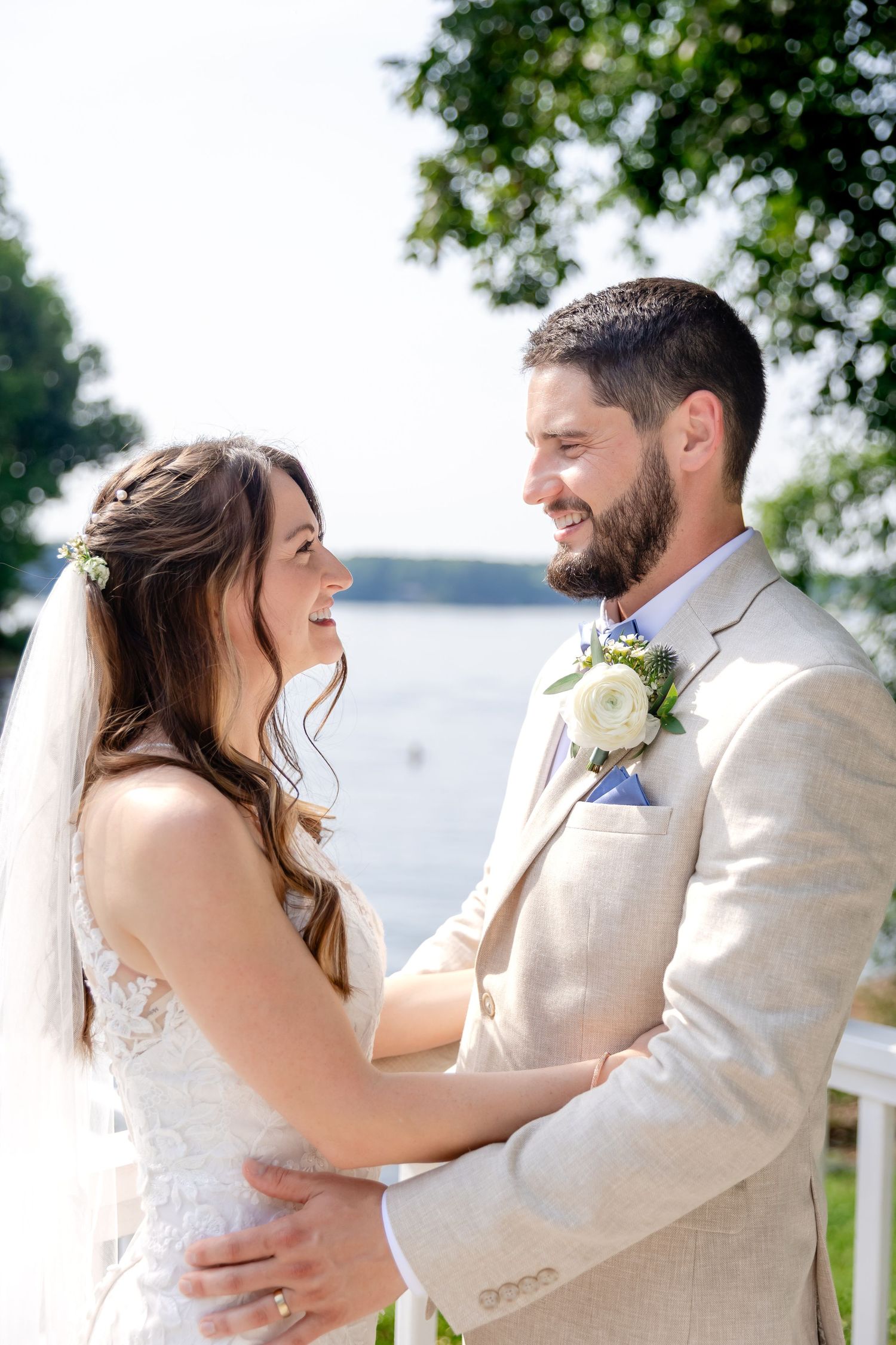 Smiling bride and groom embrace outdoors near a lake, she in lace gown with veil, he in tan suit.