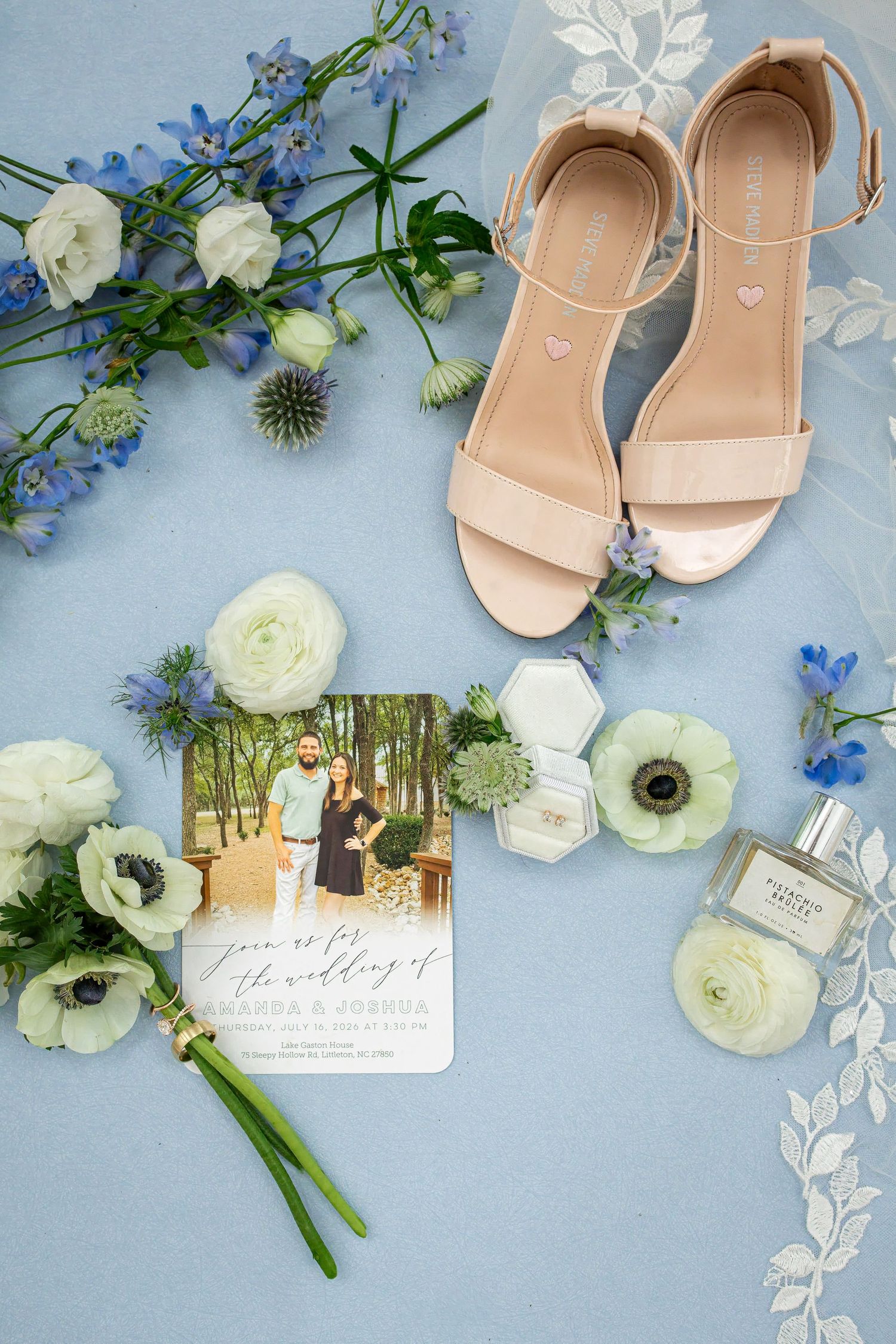 Wedding flat lay with nude sandals, blue and white flowers, invitation, ring, and perfume on blue background.