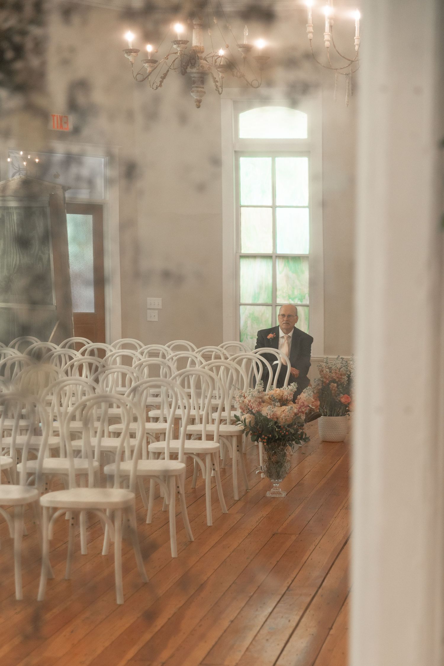 An older man in a suit sits alone among rows of white chairs in a sunlit wedding venue with a chandelier and flowers.