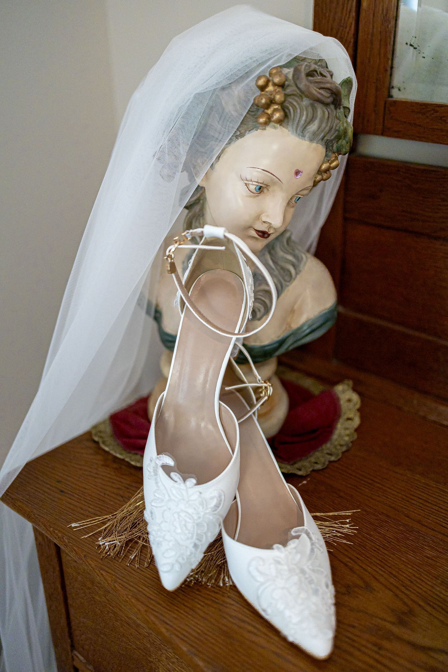 White lace bridal shoes with ankle straps rest on a wooden dresser beneath a sheer veil draped over a vintage porcelain bust.