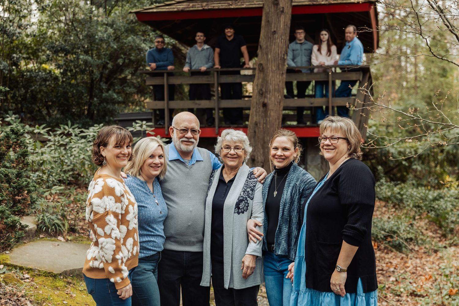 Large family group portrait outdoors with some members on a wooden deck and others standing below among trees.