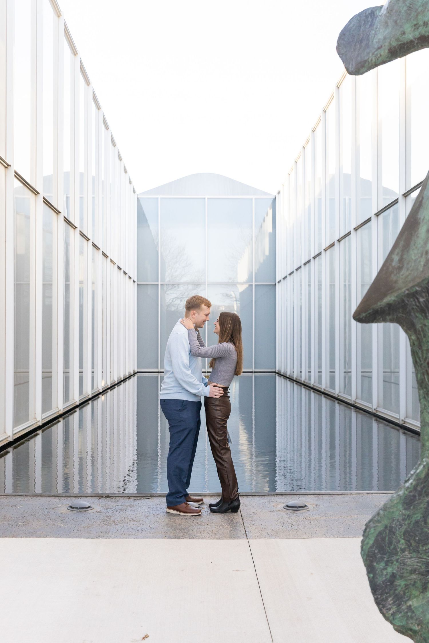 Couple facing each other between reflective modern architecture walls with a bronze sculpture nearby.