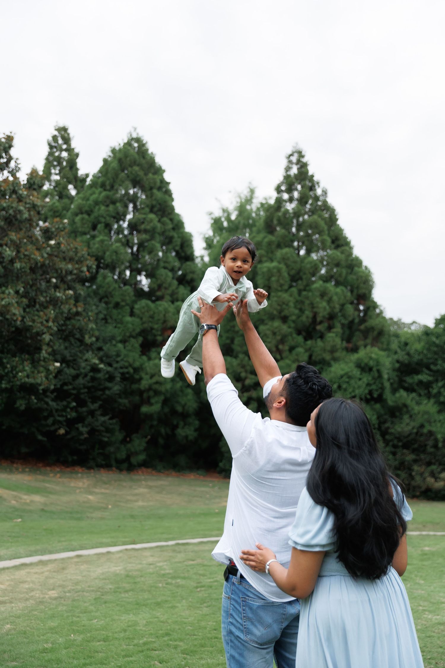 A father lifts a smiling toddler high into the air while the mother watches in a grassy park with tall evergreen trees.