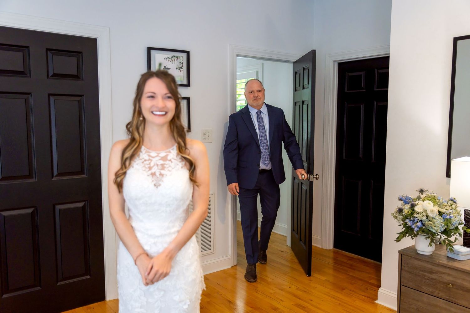 Smiling bride in a white lace wedding dress stands in a bright hallway as a man in a navy suit walks through a doorway behind her.