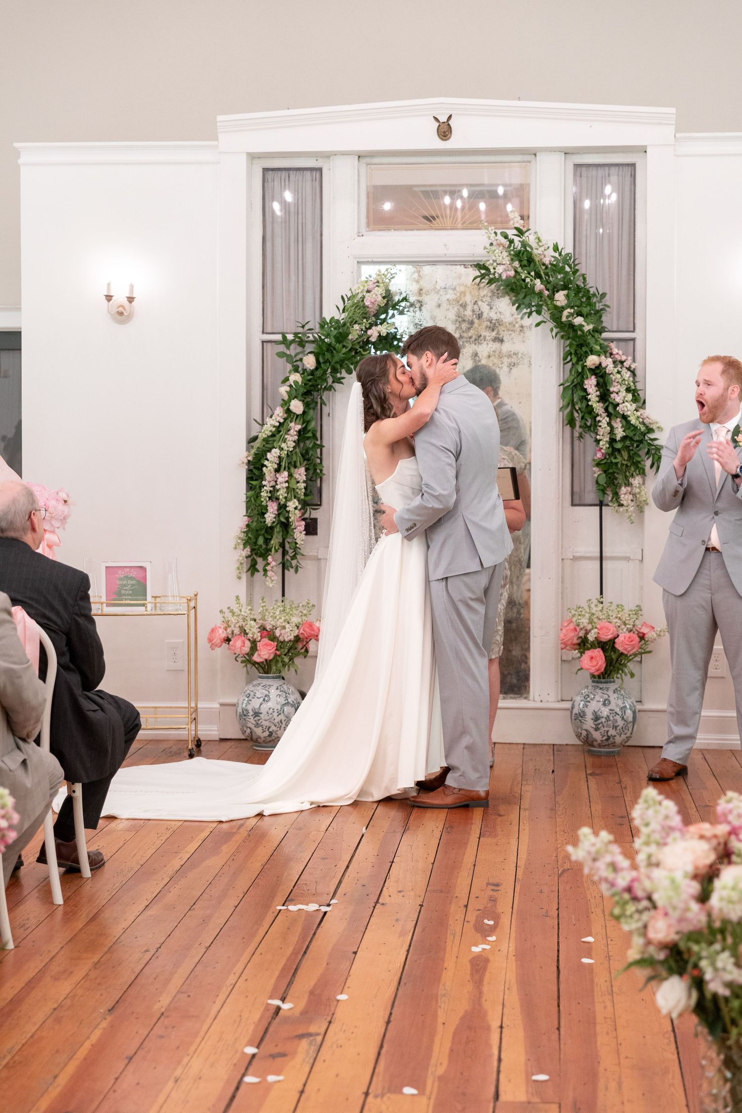 Bride in a white gown and groom in a gray suit share a kiss at their wedding ceremony beneath a floral greenery arch as a guest cheers.