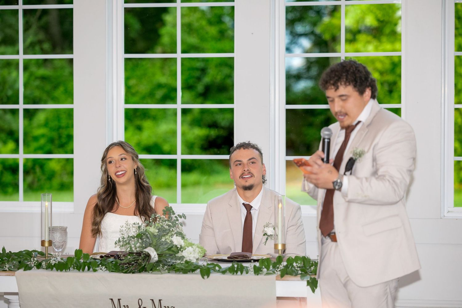 Groomsman in a beige suit gives a wedding toast with a microphone as the bride and groom smile at the decorated head table.