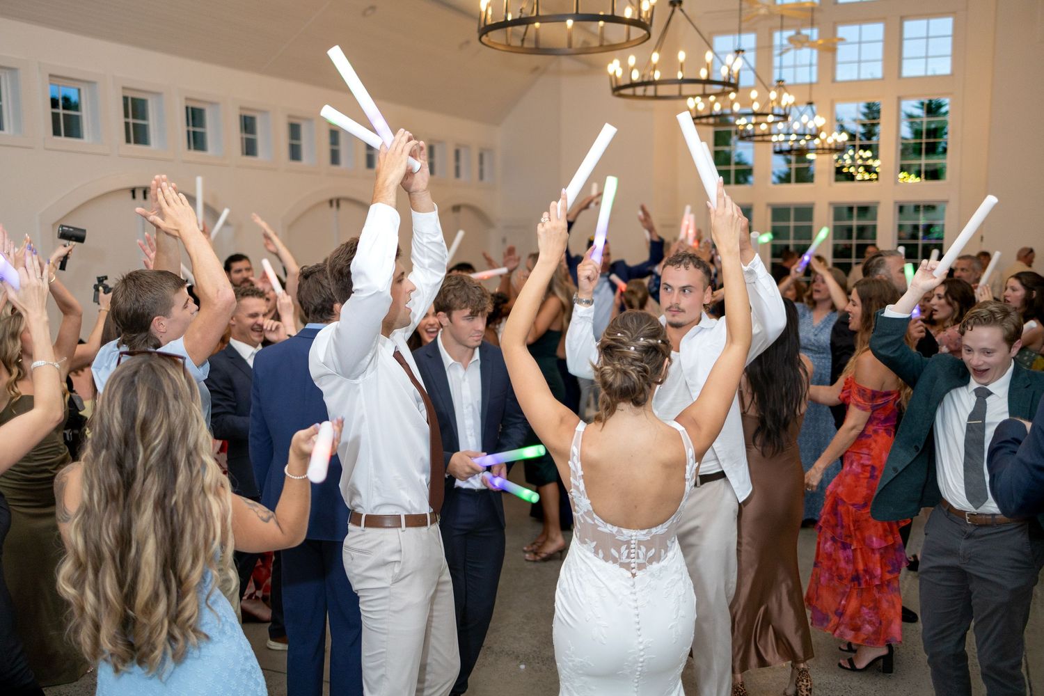 Bride in a white gown and wedding guests dance with glow sticks raised high in a bright reception hall with chandeliers.