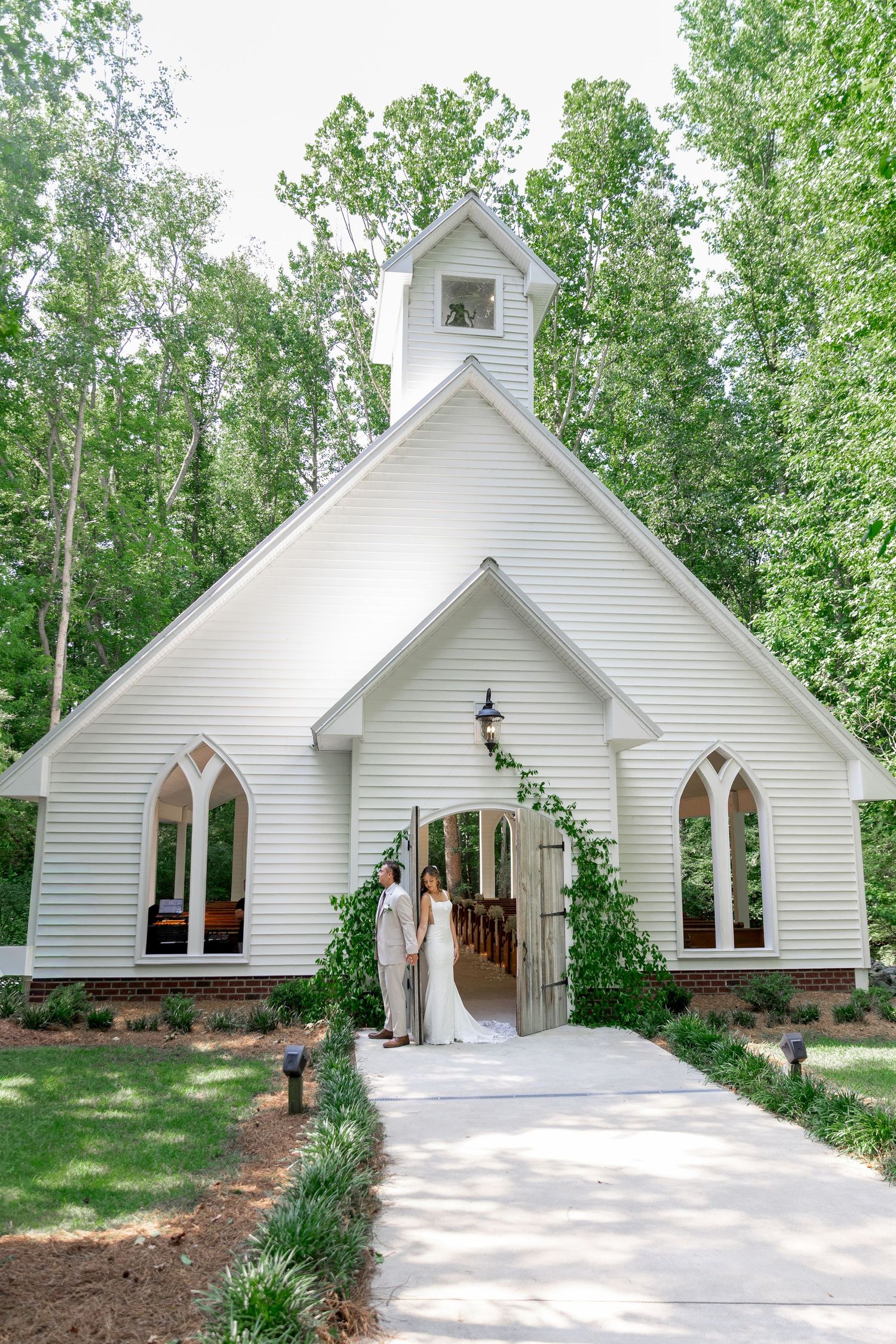 Bride and groom pose together at the entrance of a charming white chapel surrounded by lush green trees.