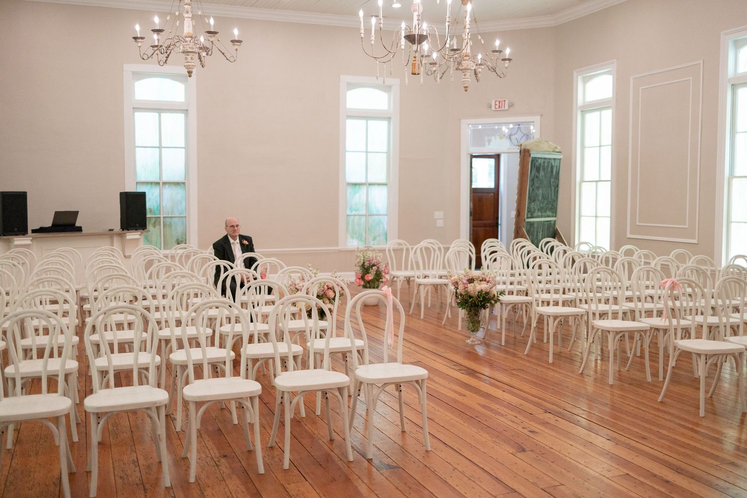 Elegant white wedding ceremony venue with rows of white chairs, chandeliers, and tall windows on hardwood floors.