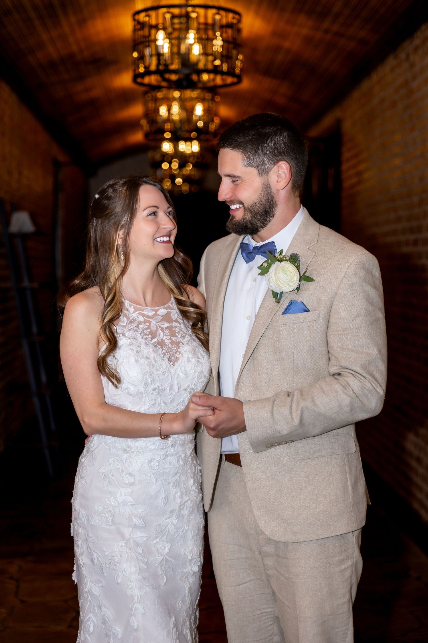 A smiling bride and groom hold hands under a chandelier in a brick hallway, wearing a white lace gown and tan suit.