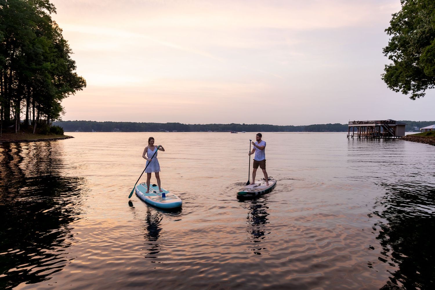 Two people stand on paddle boards gliding across calm lake water at sunset, with a wooded shoreline and a dock in the distance.