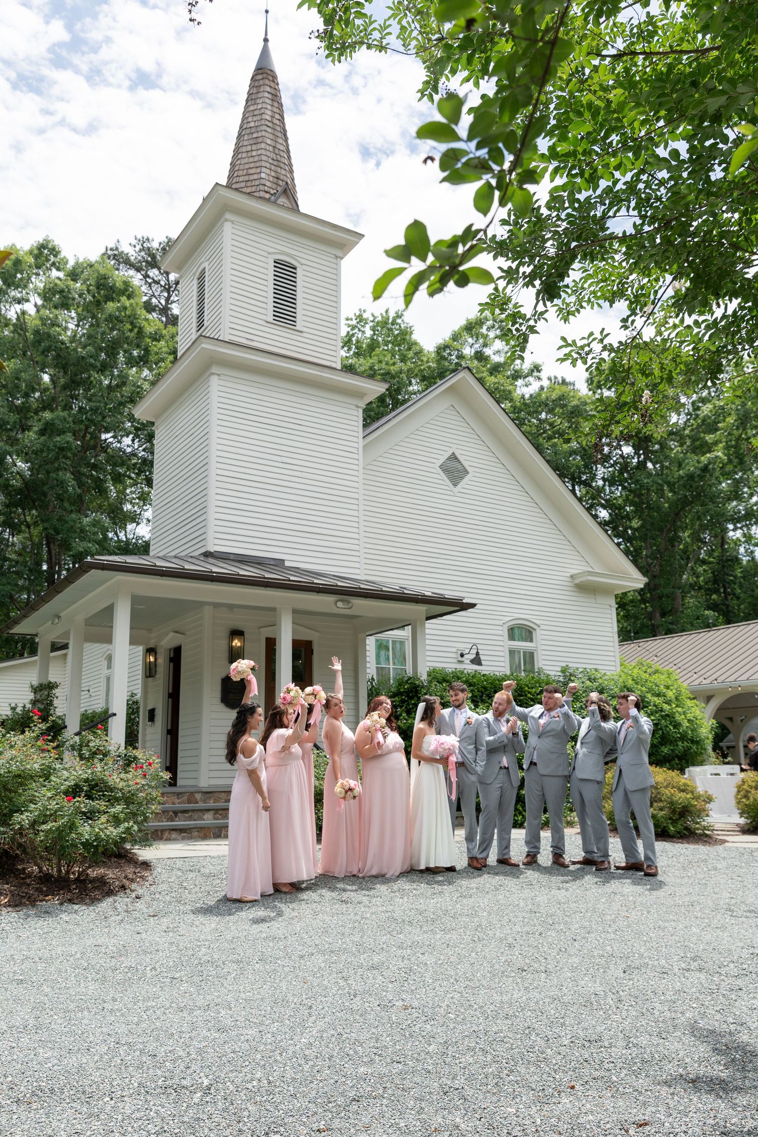Bridesmaids in blush dresses and groomsmen in gray suits cheer outside a white wooden chapel with a steeple at a summer wedding.