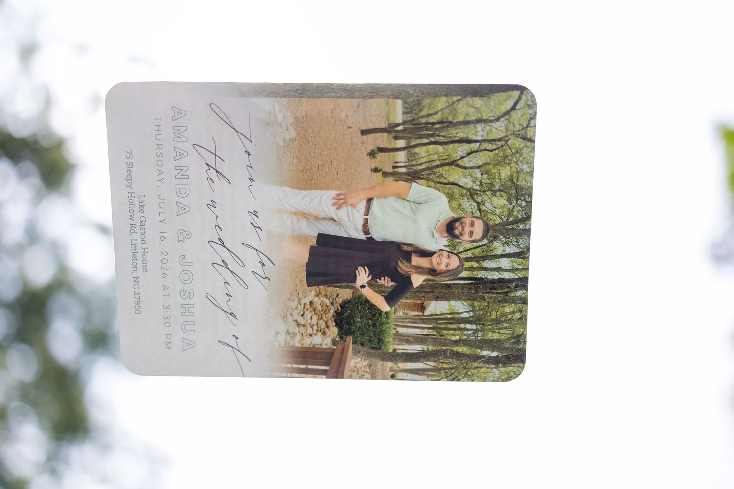 A wedding invitation card featuring a photo of the couple held up against a blurred outdoor background.