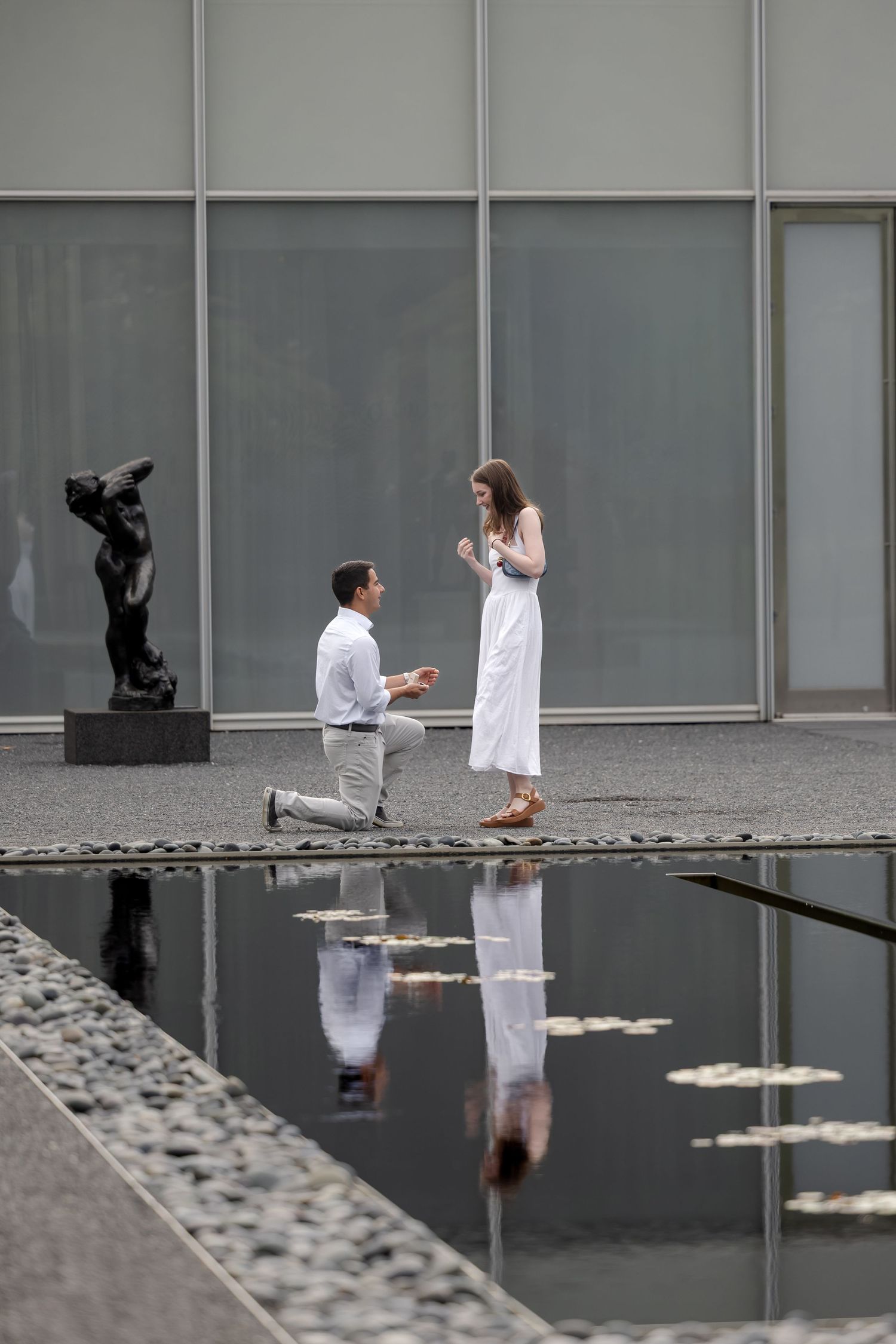 Man proposes on one knee to woman in white dress beside reflective pool with sculpture in background.
