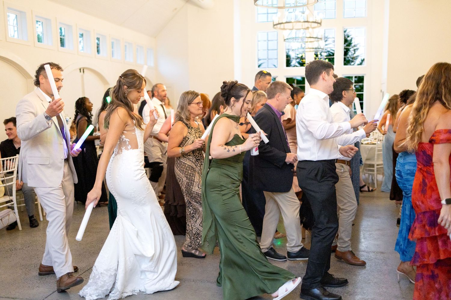 Bride in a white gown dances with wedding guests in elegant attire inside a bright reception hall with tall windows and a chandelier.