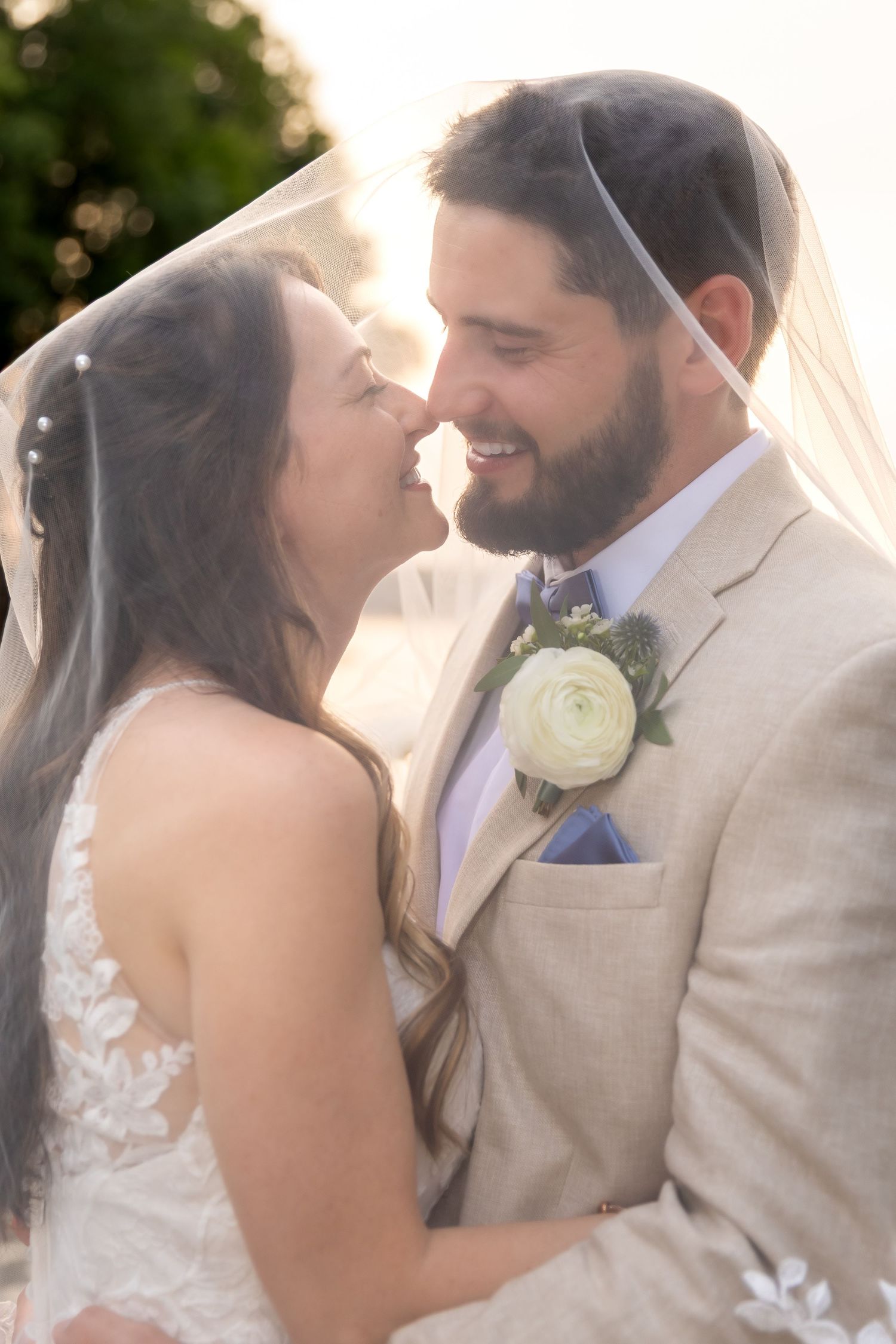 Bride and groom share a romantic moment under a veil, smiling closely, groom in tan suit with white boutonniere.