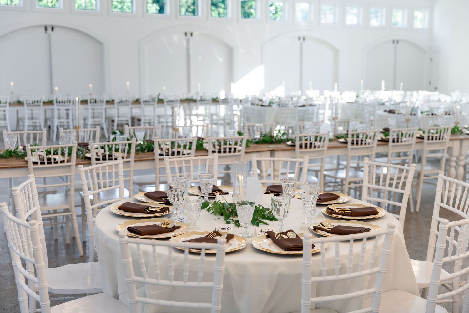 White wedding reception venue with chiavari chairs, round tables, and warm wood charger plates.