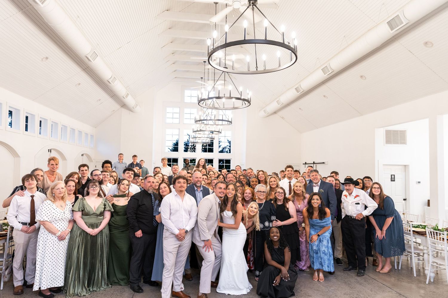 Large wedding party and guests pose together in a bright reception hall with chandeliers, the bride in a white gown at the center.