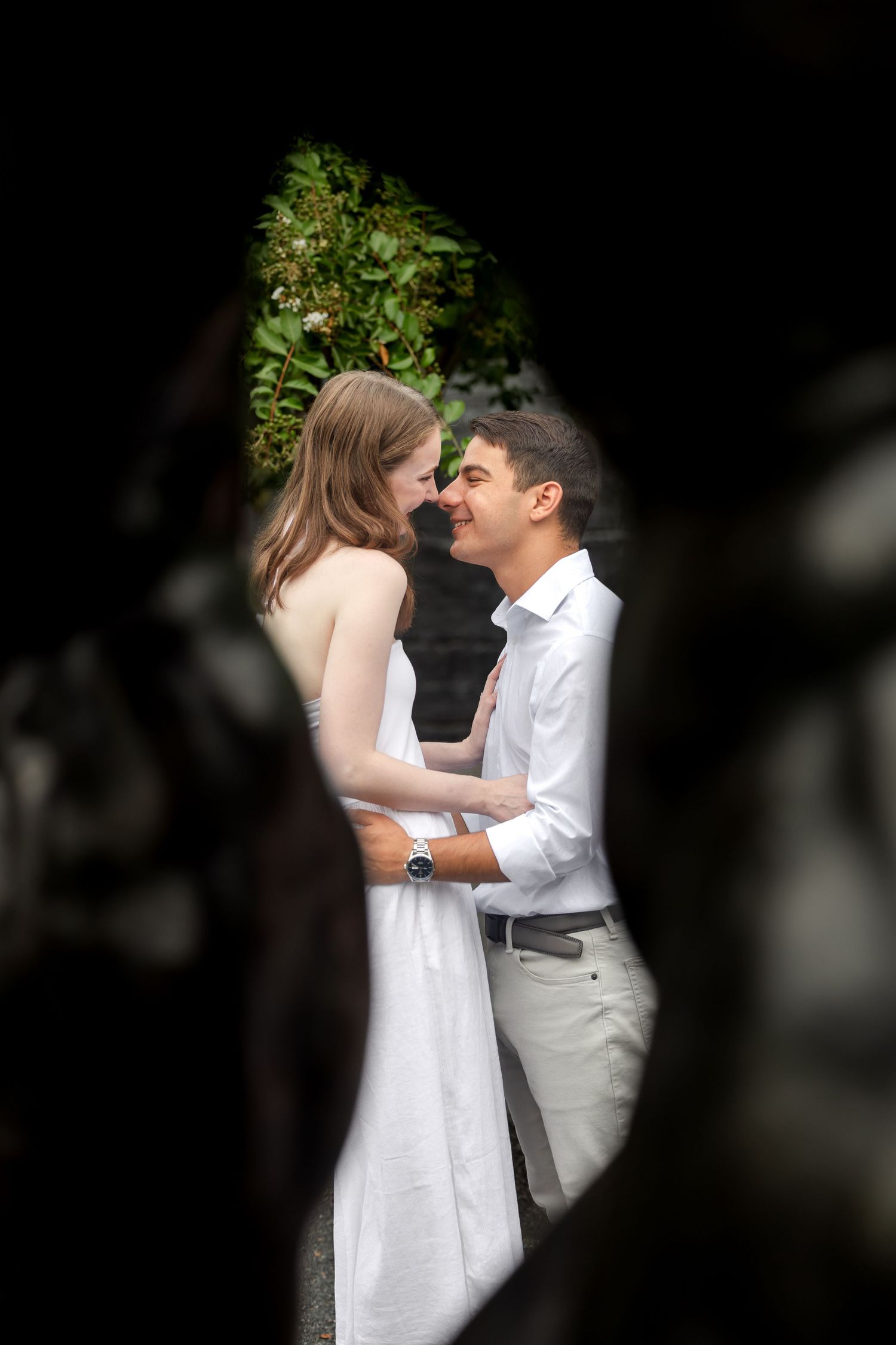 Couple in romantic embrace outdoors, woman in white dress and man in white shirt, framed by dark foliage.