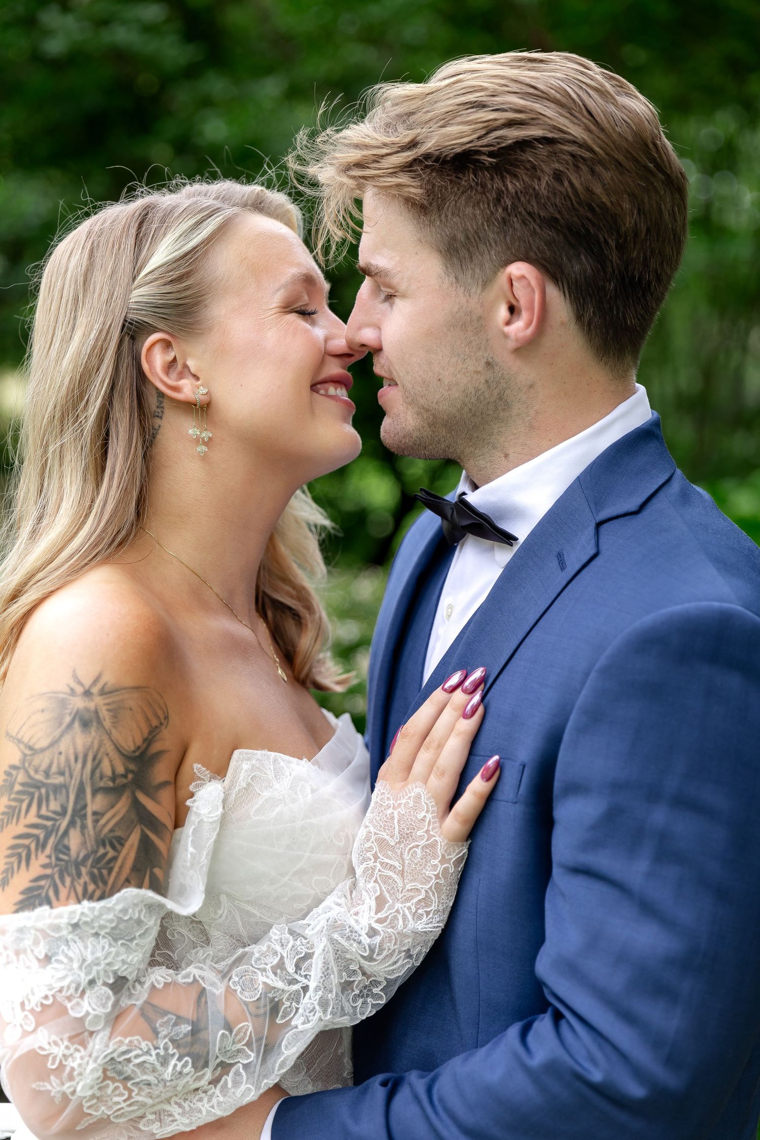 A smiling bride and groom in a blue suit share a romantic moment outdoors, nearly kissing amid lush green foliage.