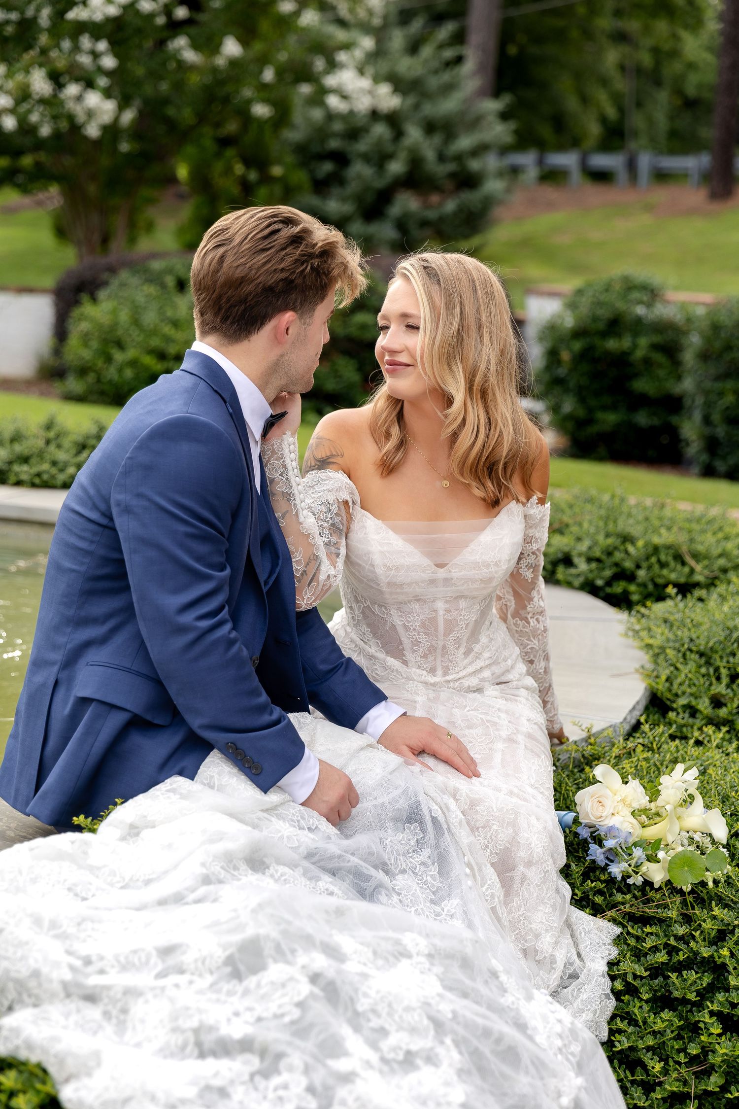A bride in lace gown and groom in blue suit share an intimate moment seated in a lush garden setting.