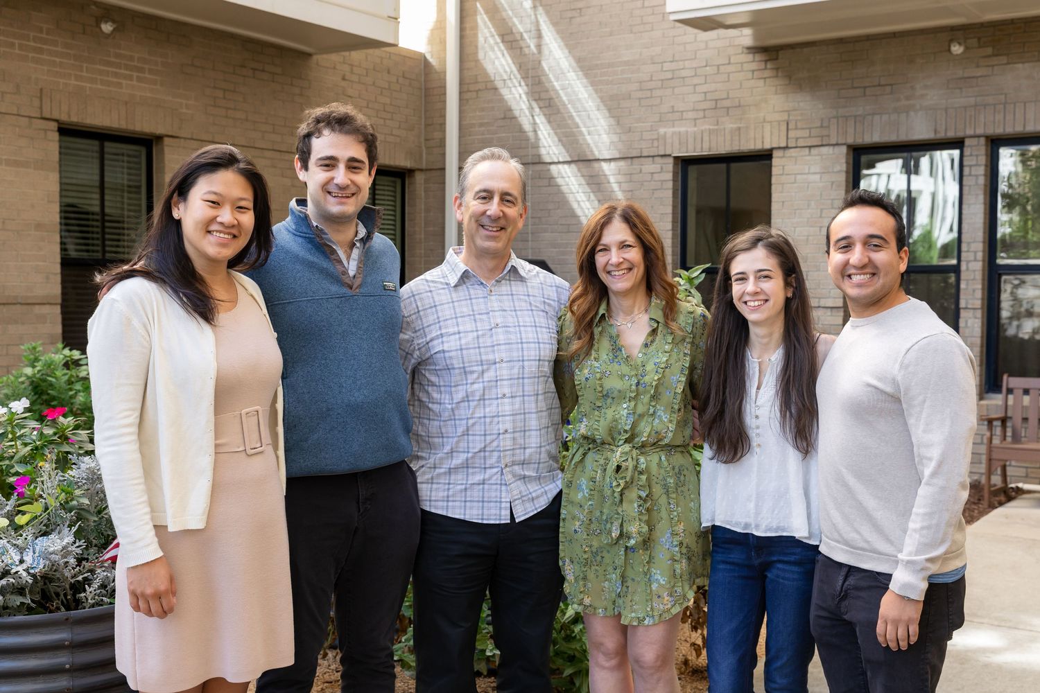 Six smiling adults stand close together in a sunlit brick courtyard, posing for a casual group photo beside green plants and large windows.