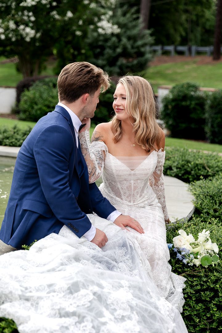 Bride in lace off-shoulder gown and groom in navy suit sit together in a lush garden setting.