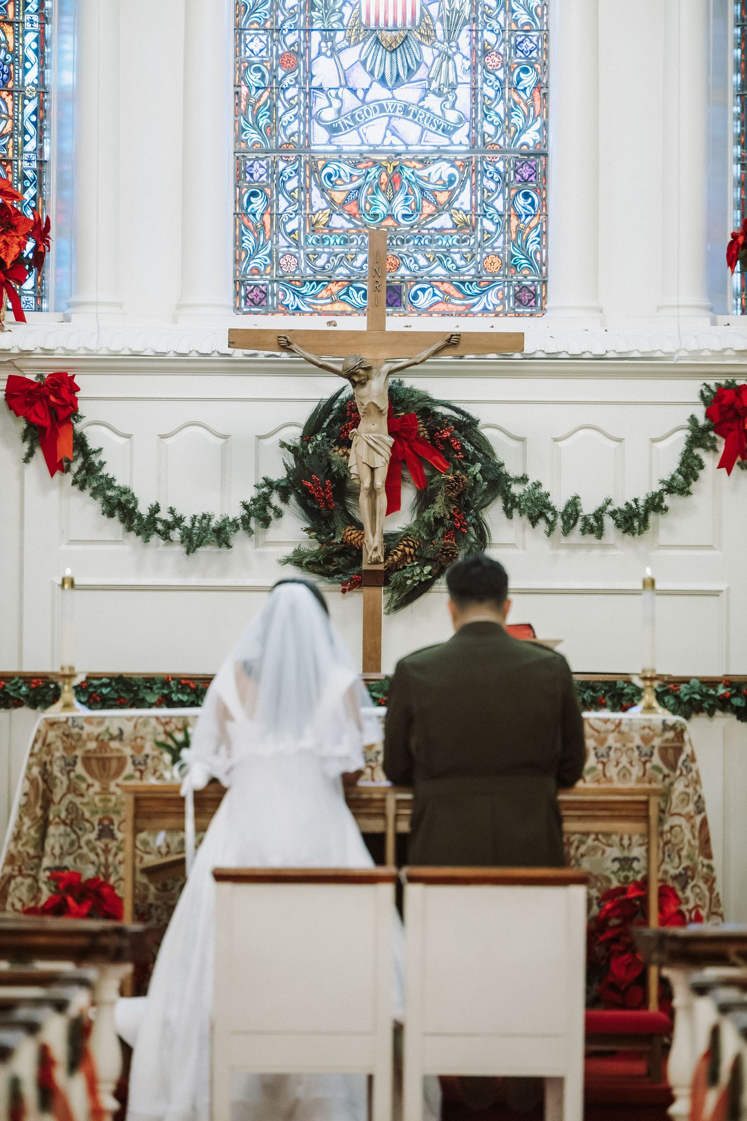 Bride in white gown and groom face altar with crucifix and Christmas wreath in decorated church.