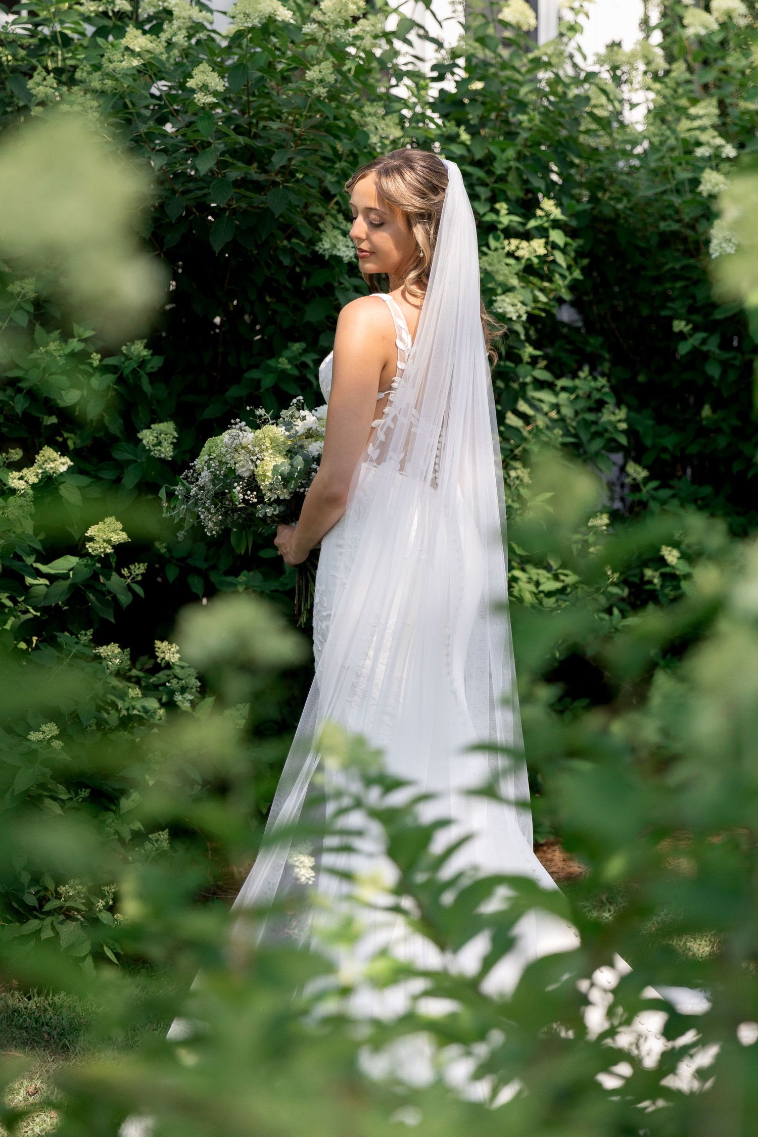 Bride in white lace gown and veil holding bouquet surrounded by lush green garden foliage.