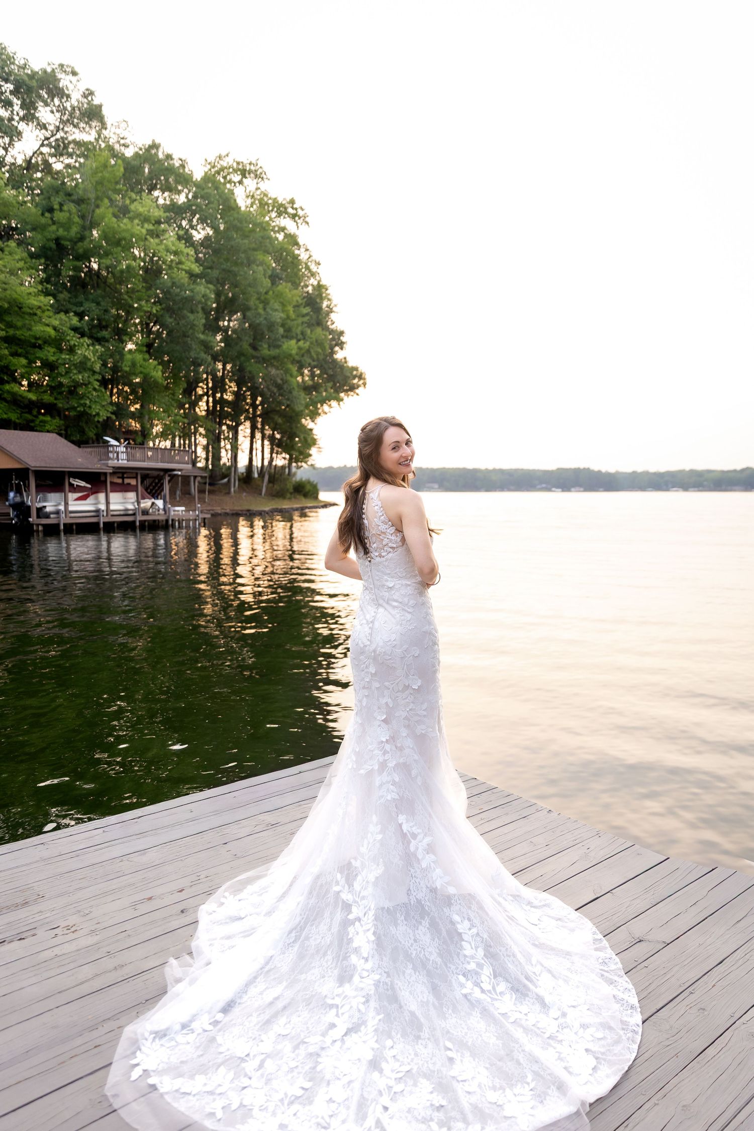 Bride in a lace mermaid wedding gown with a long train looks back over her shoulder on a wooden lakeside dock at sunset.