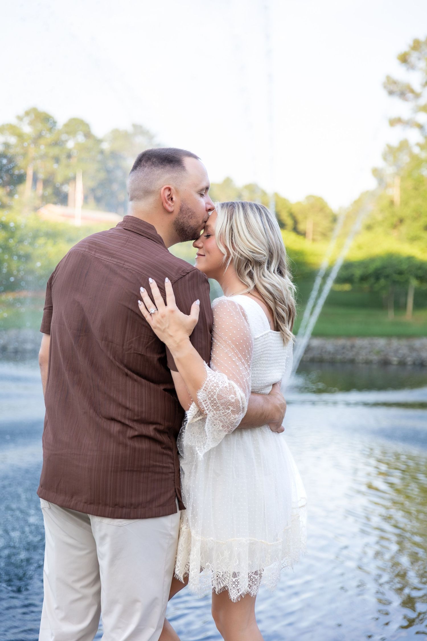Couple embracing by a fountain pond as he kisses her forehead, her hand with a ring resting on his shoulder, in a sunny park.