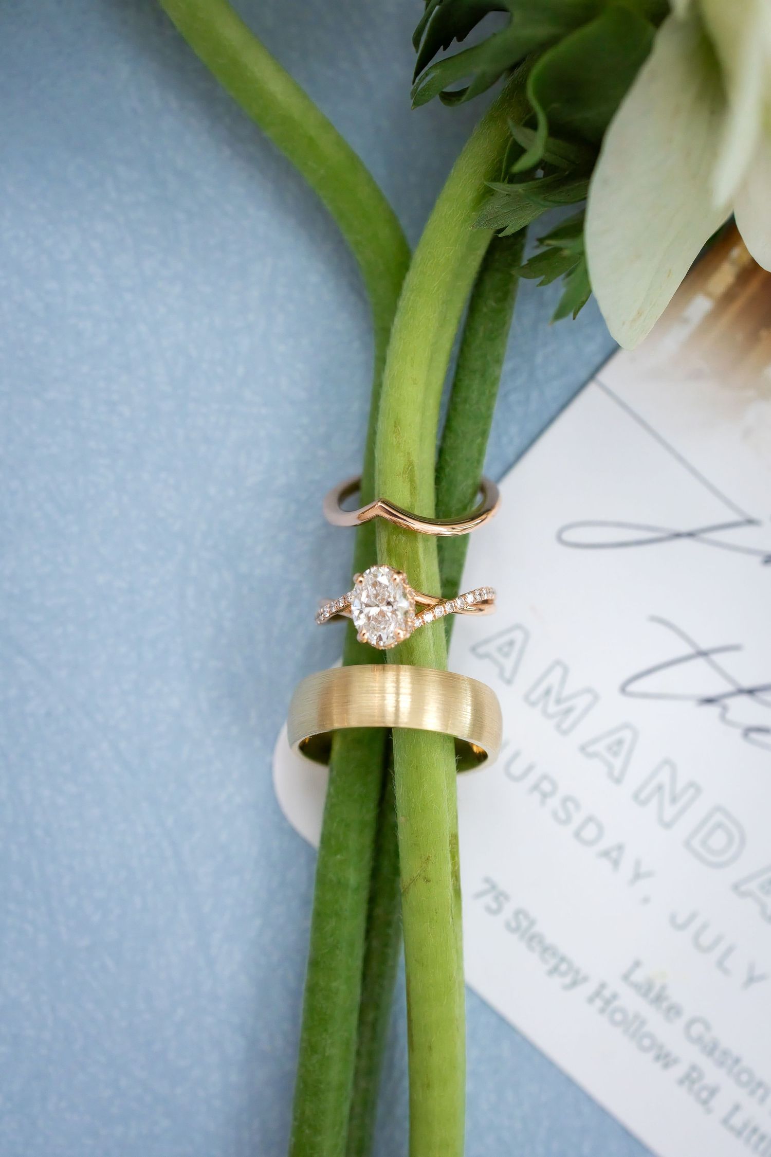 Rose gold oval diamond engagement ring, chevron band, and gold wedding band displayed on green flower stems.